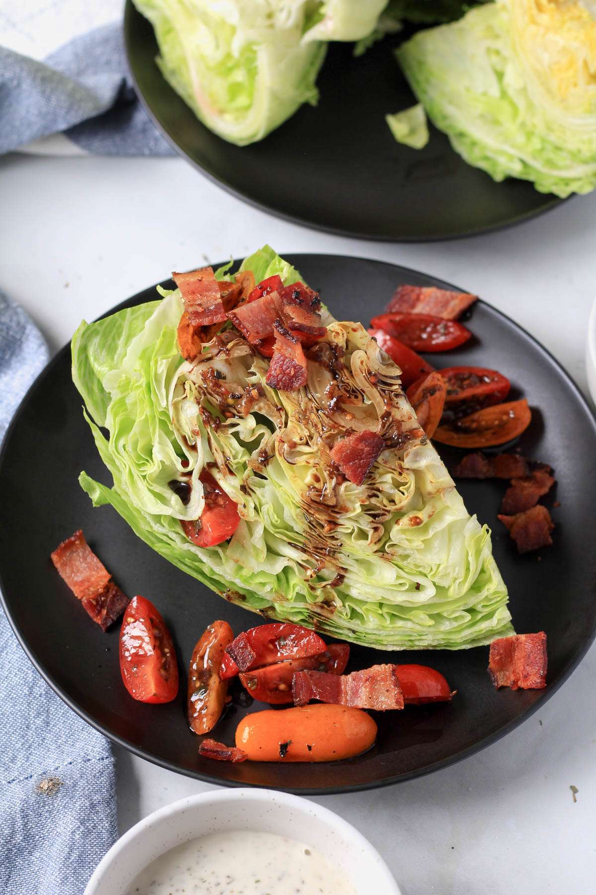 A black plate with a quarter of iceberg lettuce topped with balsamic glazed tomatoes and topped with cooked bacon.