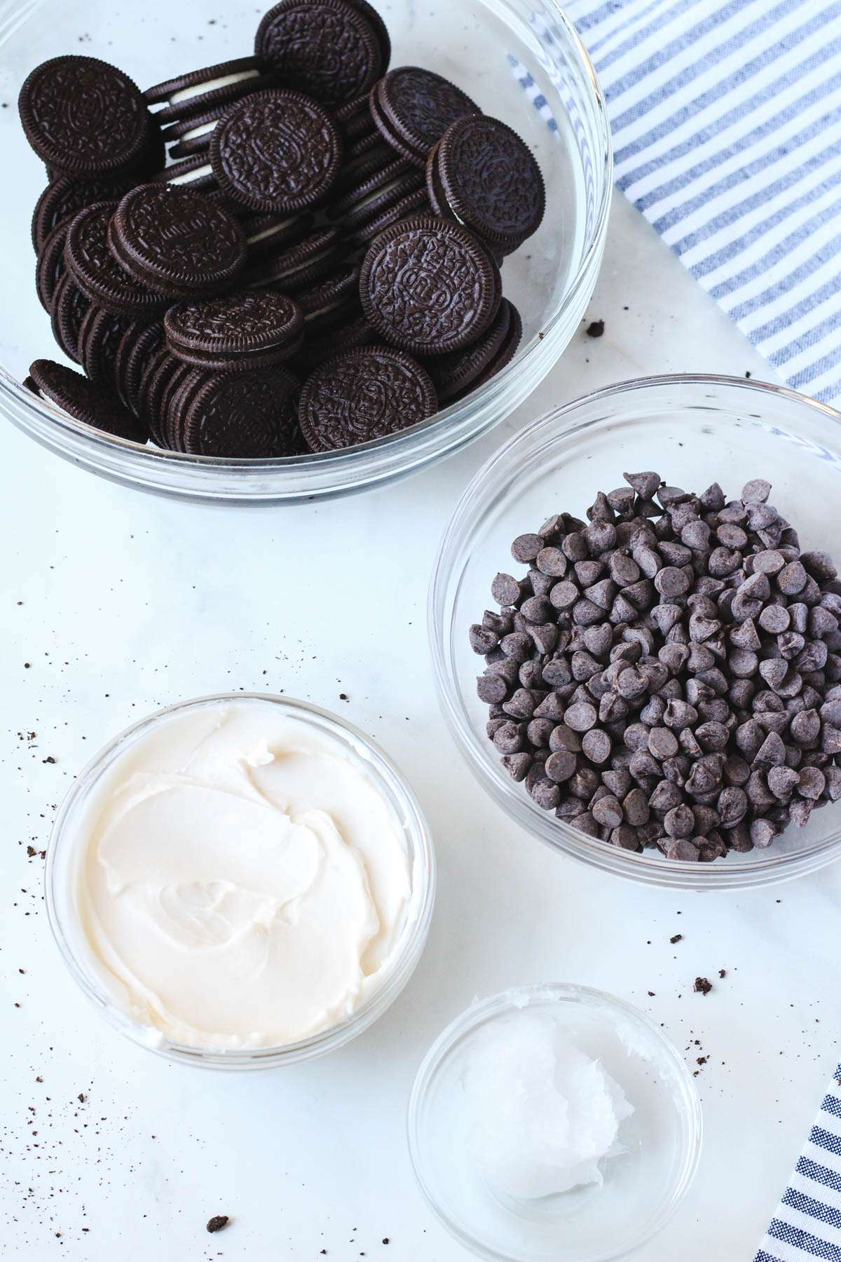 Ingredients for Vegan Oreo Truffle Balls in glass bowls on a white counter with a blue and white striped dish towel to the right.