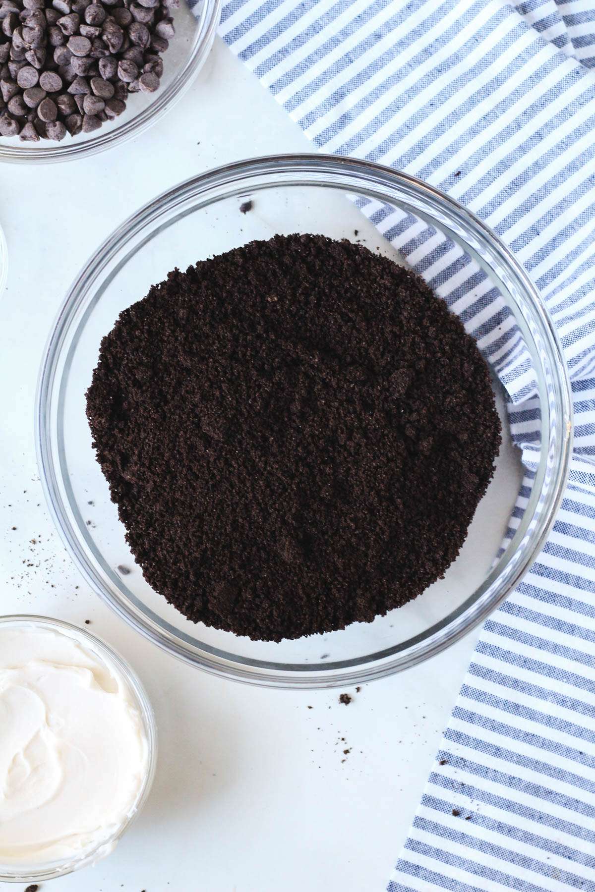 A glass mixing bowl with oreo cookie crumbs in the middle and a small bowl of oreo cookie crumbs to the bottom left.