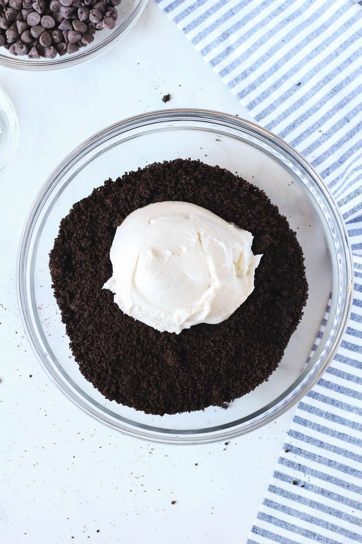 A glass mixing bowl on a white counter with oreo cookie crumbs topped with vegan cream cheese before mixing together.