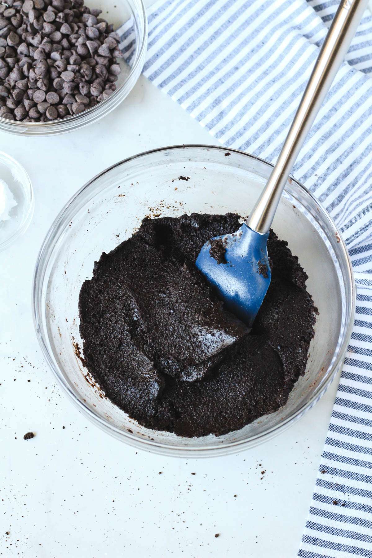 A glass bowl with a blue and silver spatula and a mixture of Oreo cookie crumbs and dairy free cream cheese.