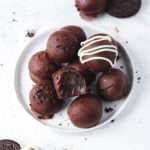 A white rimmed plate of vegan Oreo truffle balls on a white counter with oreo cookies and crumbles around the plate.