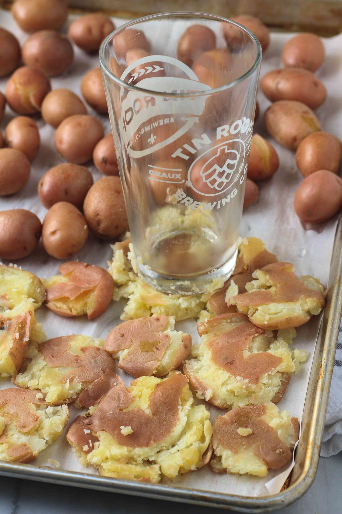 A rimmed baking sheet lined with parchment paper and topped with small potatoes with a pint glass smashing the potatoes.