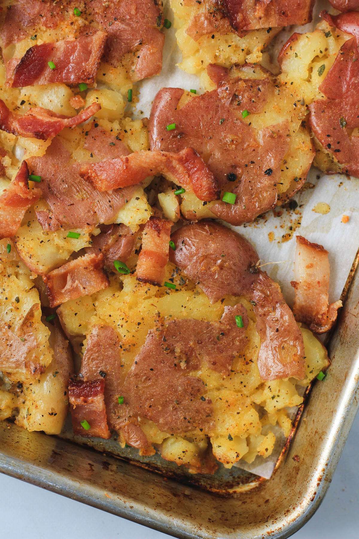 The bottom right corner of a rimmed baking sheet with smashed potatoes topped with bacon and chive.