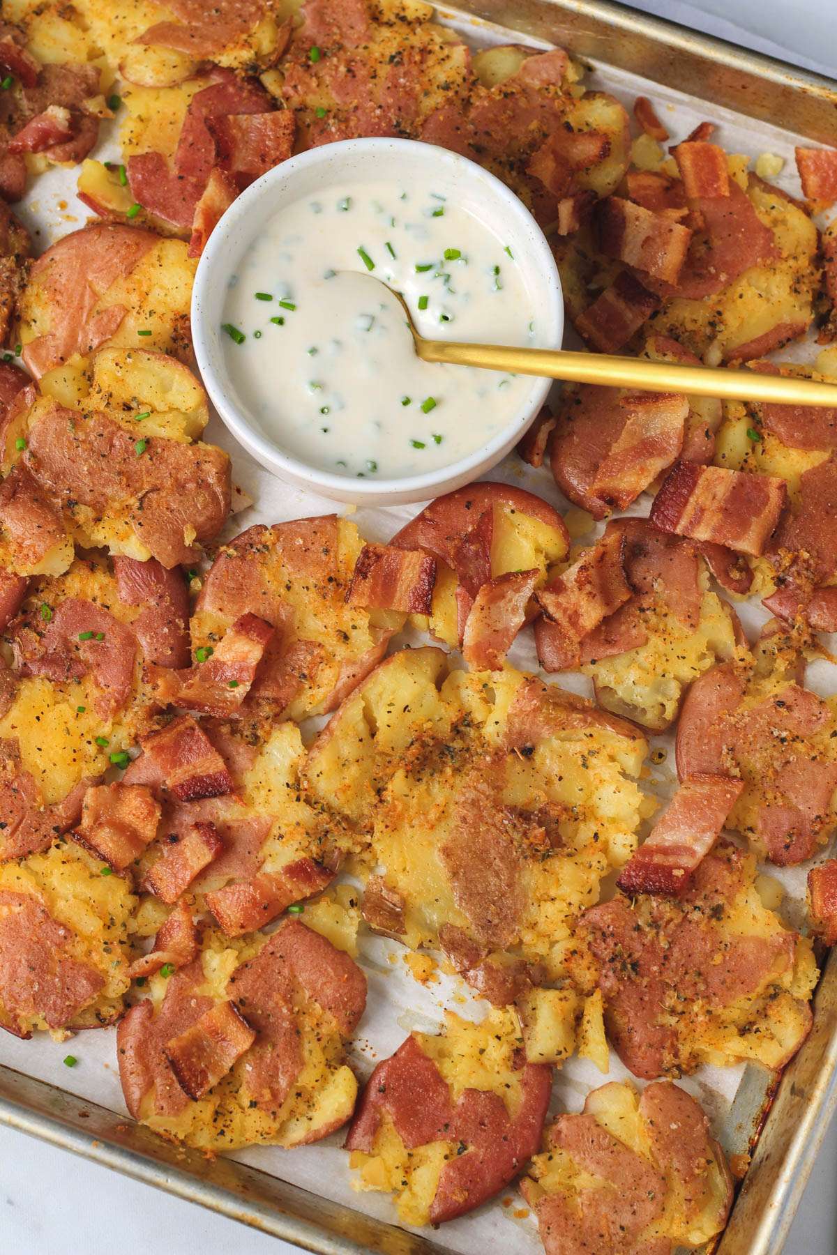 A sheet pan with breakfast smashed potatoes topped with bacon bits and a bowl of chive sour cream with a gold spoon at the top of the baking sheet.