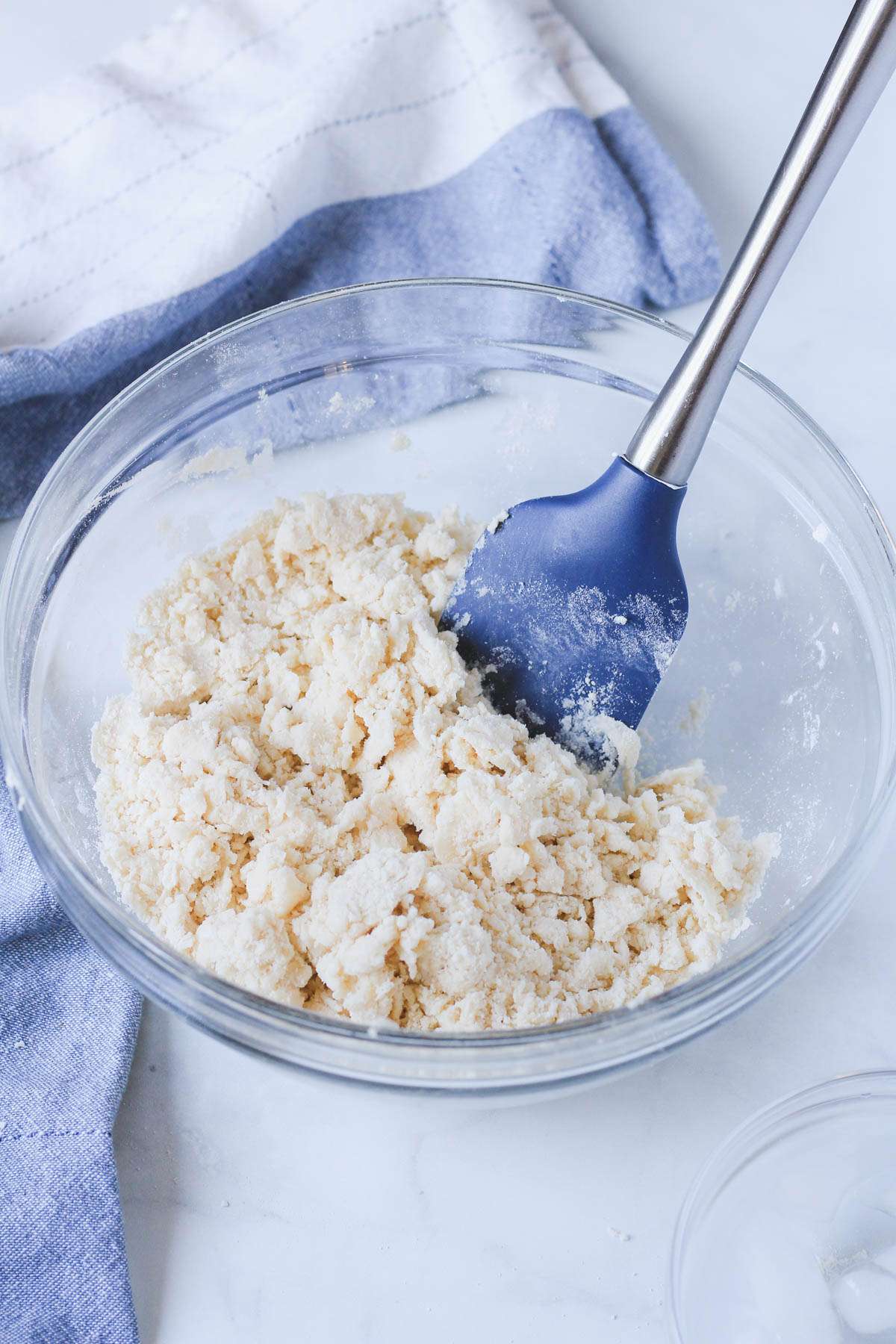 A blue rubber spatula folding ice water into a dough in a glass bowl.
