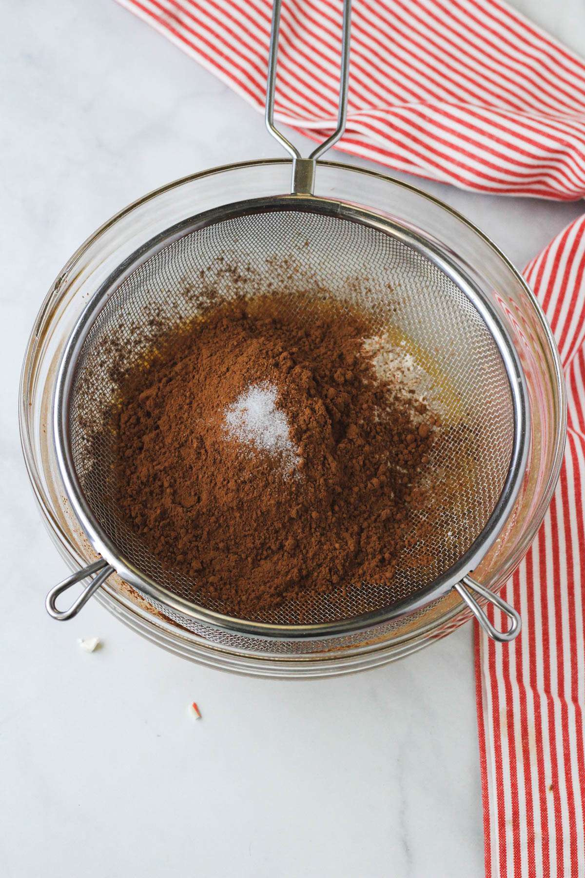 A fine mesh sieve over a glass mixing bowl with the dry ingredients being sifted into a glass mixing bowl on a white counter.