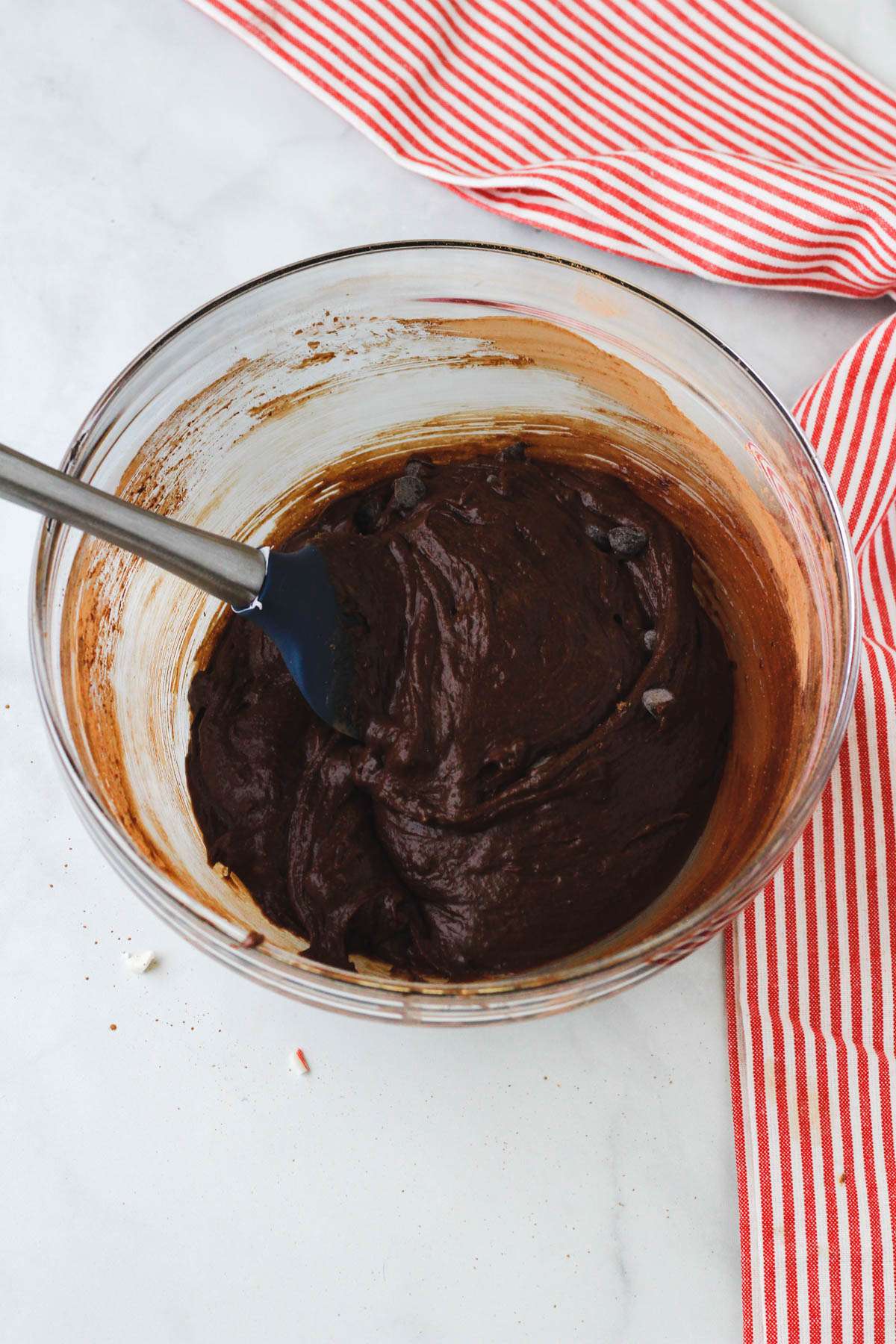 A glass mixing bowl with peppermint brownie batter and a spatula folding in the ingredients on a white counter.