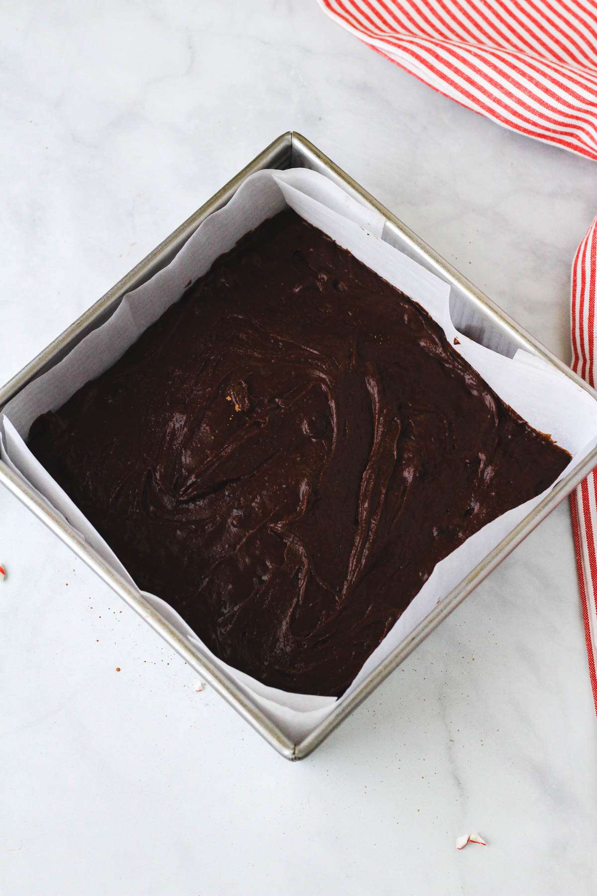 A square baking pan lined with parchment paper and filled with unbaked brownie batter on a white counter with a red and white striped towel to the right.