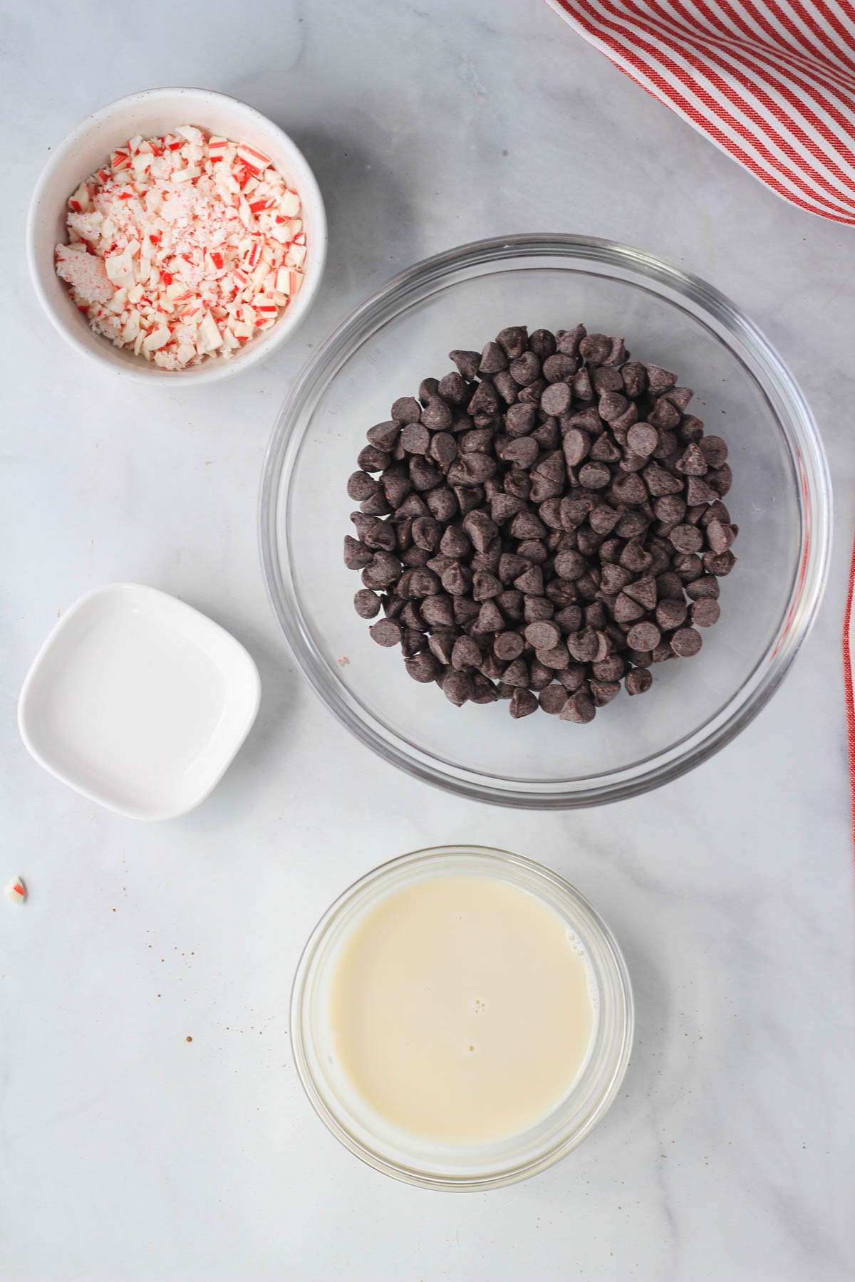 Ingredients for mint chocolate ganache in mixing bowls on a white counter with red and white striped towel to the right.