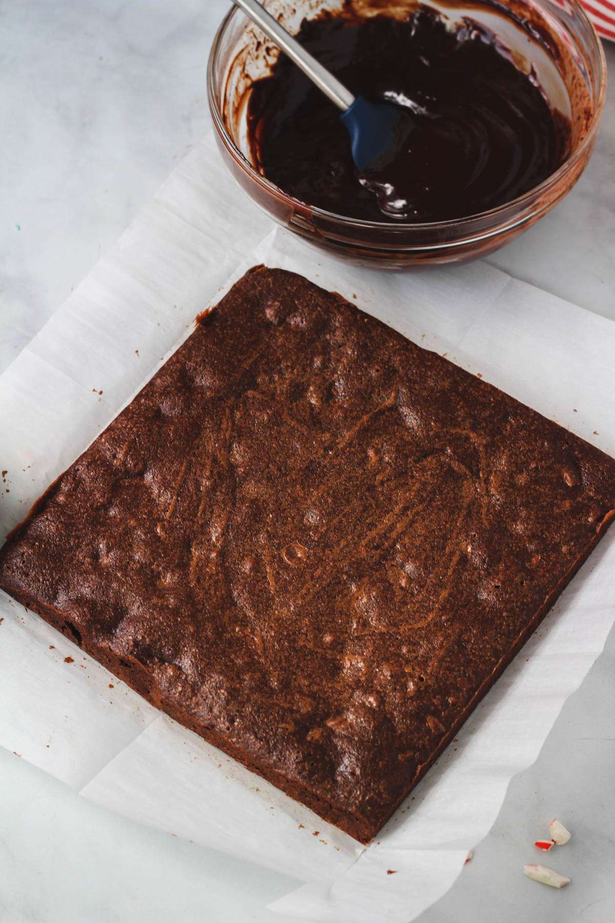 A piece of parchment paper with the baked peppermint brownie and a bowl of mint chocolate ganache in the back.