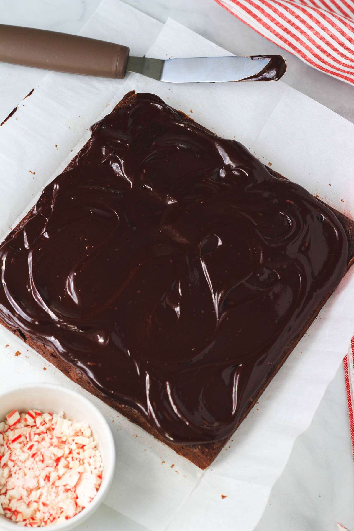 Peppermint brownies topped with mint chocolate ganache on parchment paper with a bowl of crushed peppermint in the front and an offset spatula in the back.