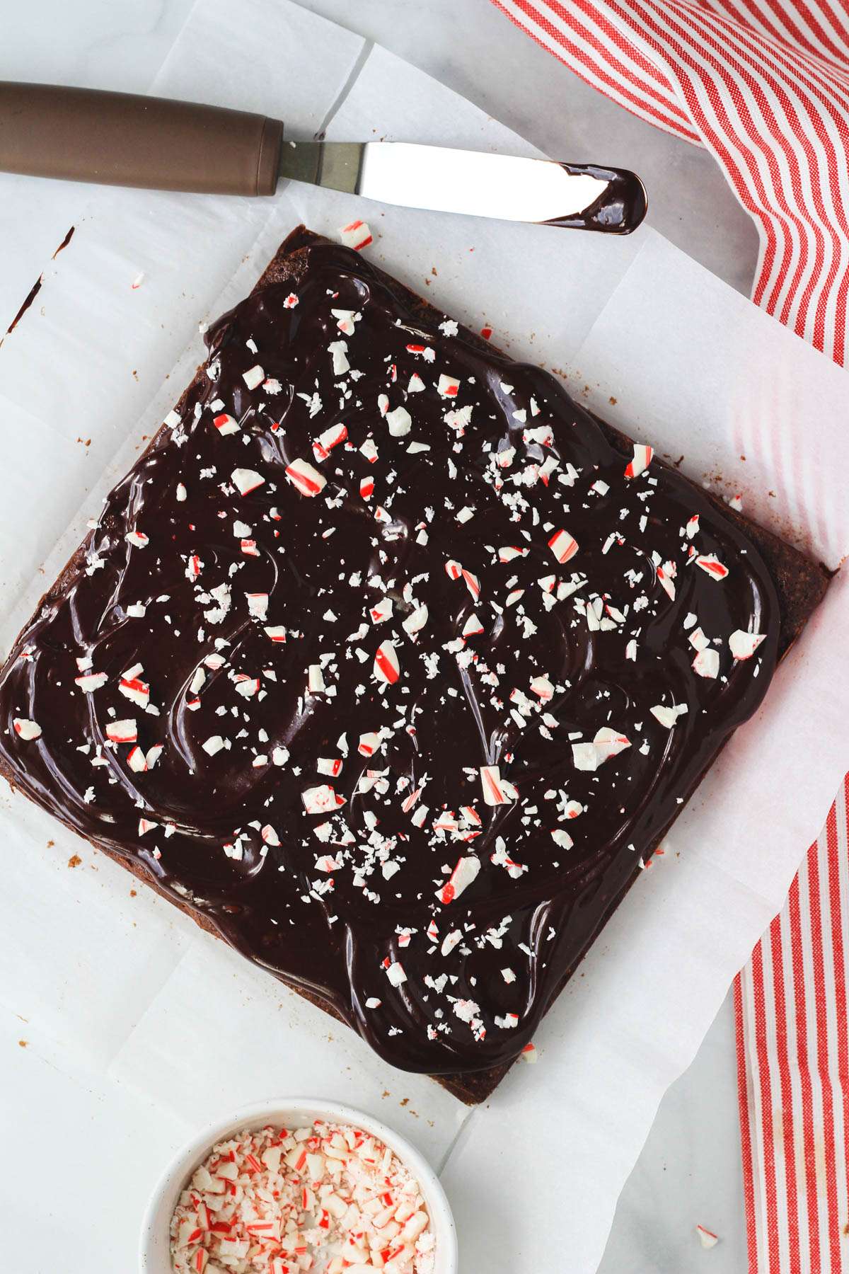 Peppermint brownies with mint chocolate ganache topped with crushed peppermint on a piece of parchment paper with a red and white striped towel to the right.