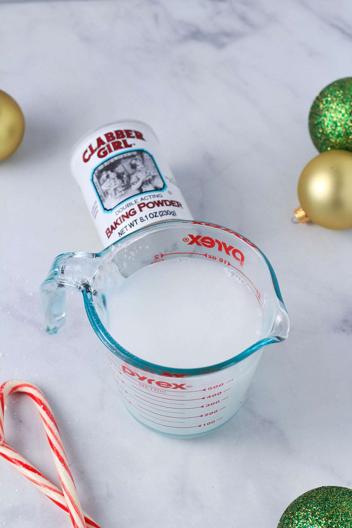 Baking powder in a liquid measuring cup with hot water on a white counter with ornaments and candy canes around the liquid measuring cup.
