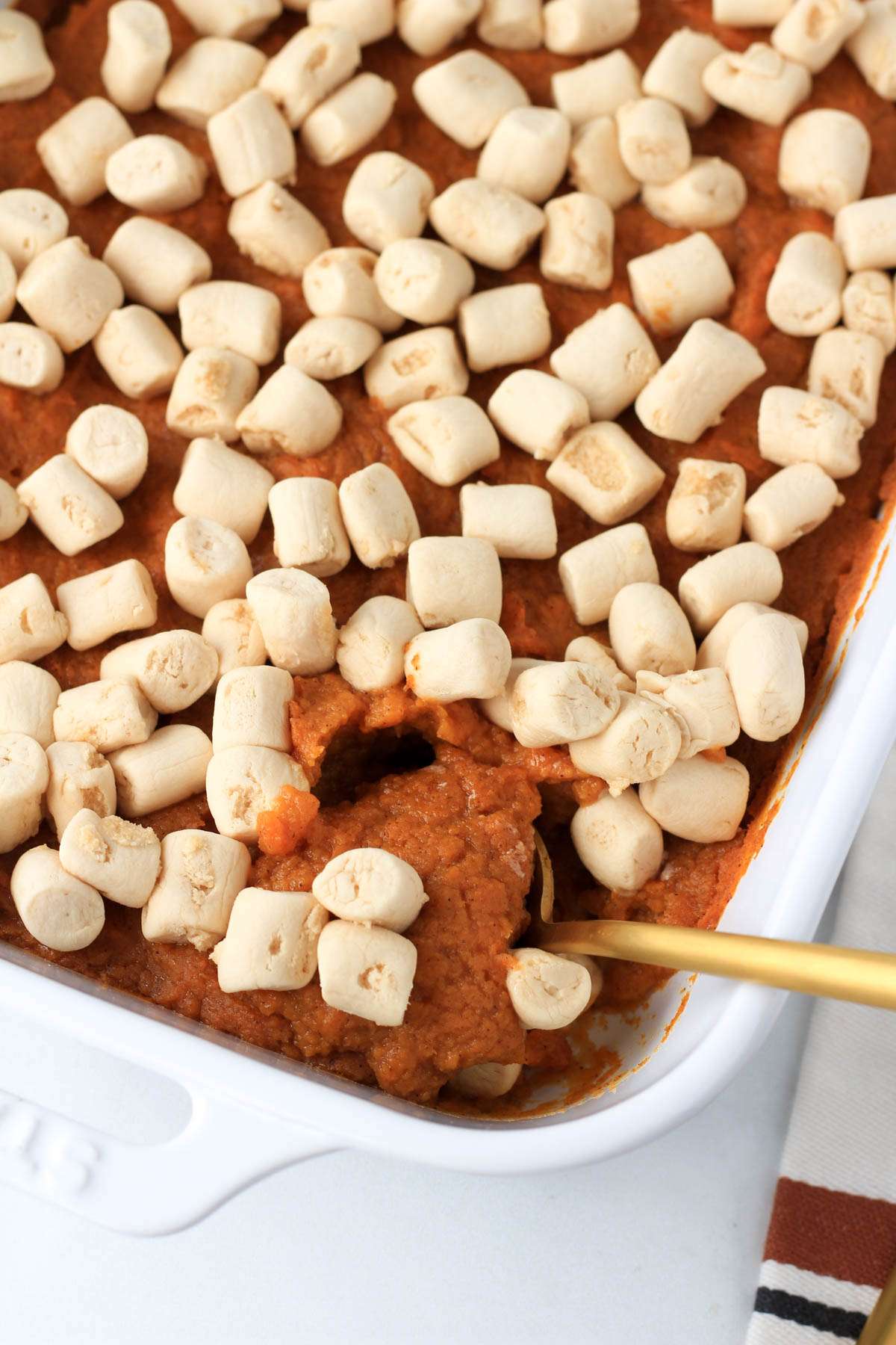 A gold spoon in a white casserole dish of maple sweet potato casserole topped with vegan marshmallows.