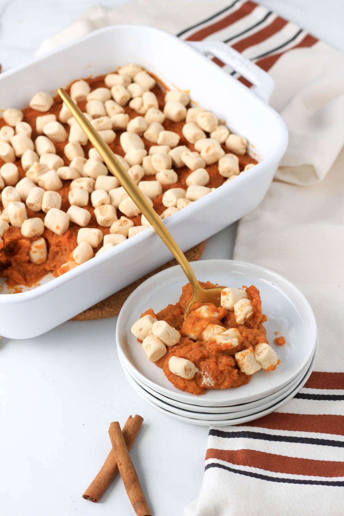 A stack of white plates topped with a scoop of sweet potato casserole and a white casserole dish in the back.