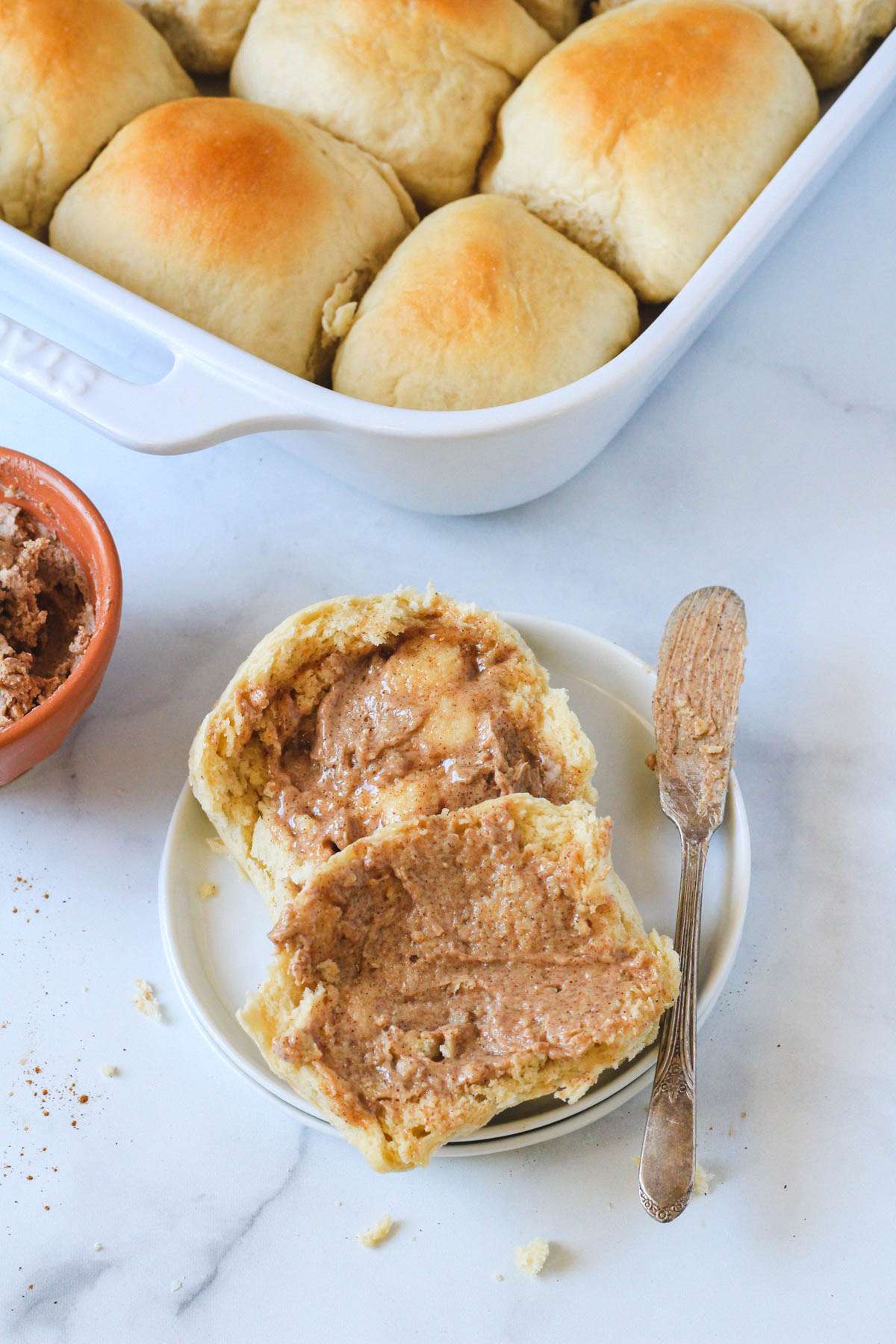 A plate with two halves of a vegan dinner roll topped with cinnamon maple butter with a pan of rolls in the back.