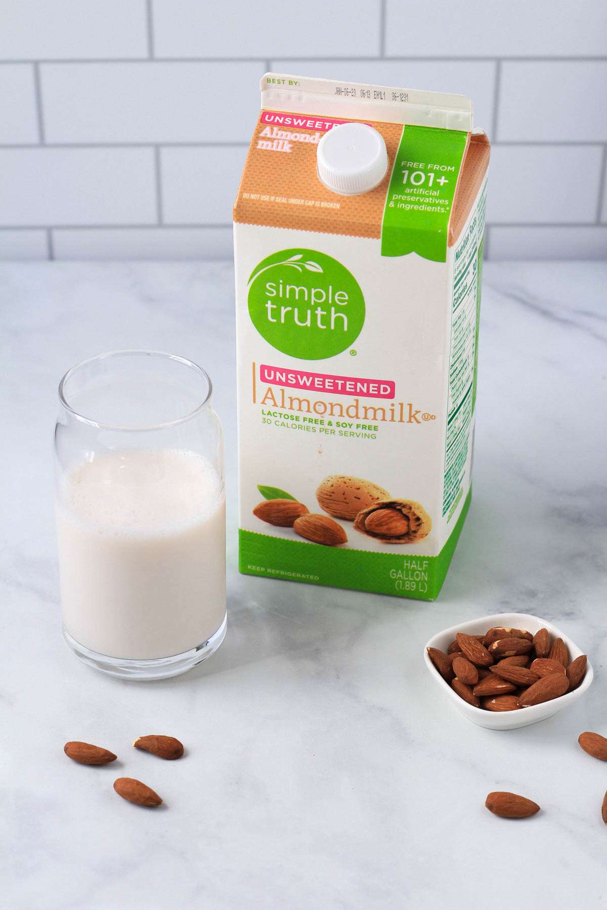 A carton of almond milk in the middle with a glass of almond milk to the left and a small dish of almonds to the right.