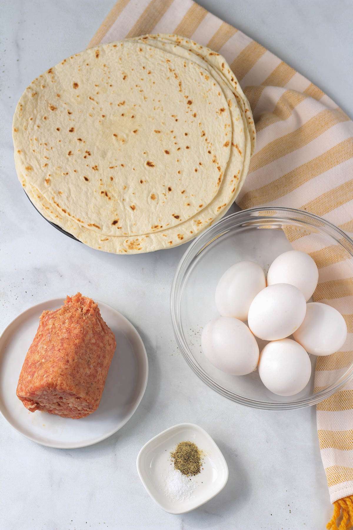 Ingredients for sausage and egg breakfast burritos on a white counter.