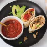 A sliced sausage and egg breakfast burrito cut it half and standing up on a black plate with a white bowl to the left filled with salsa.