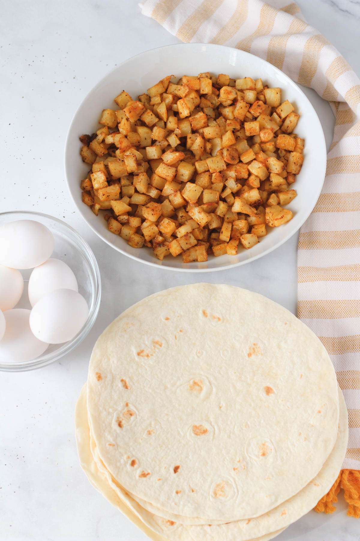 The ingredients for freezer breakfast burritos with potato and egg on a white counter.