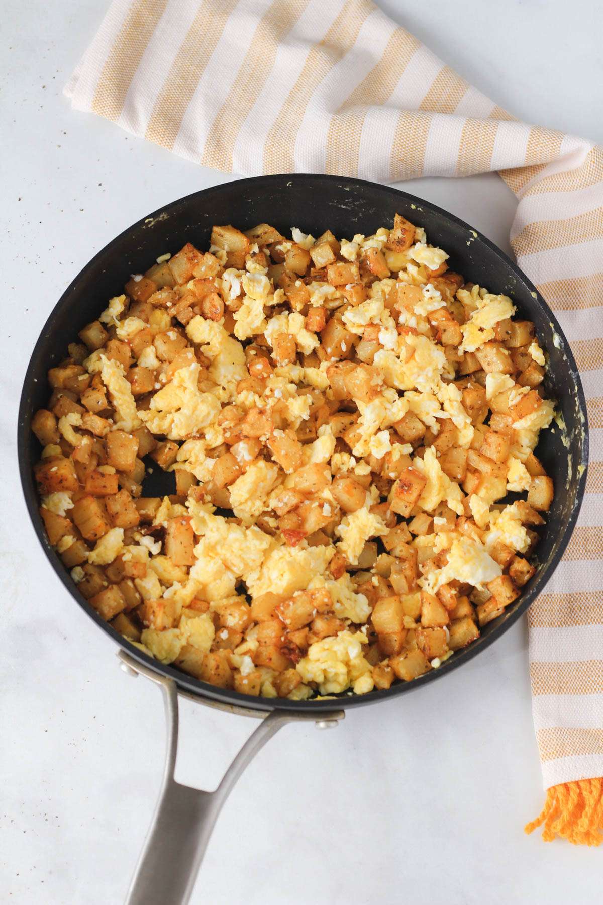 A non-stick skillet with a mixture of potato and egg next to a white and yellow dish towel.