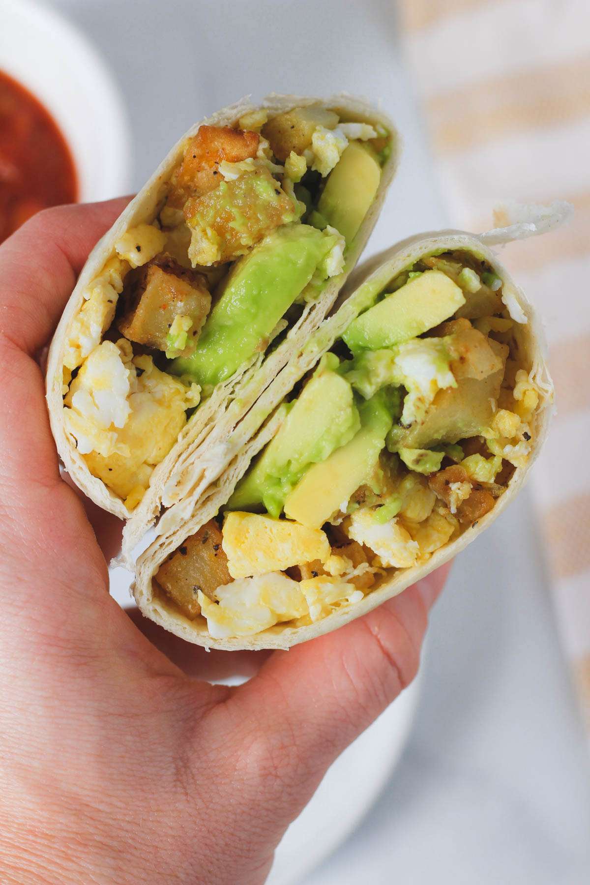 A hand holding a sliced breakfast burrito filled with egg and potato and fresh sliced avocado.