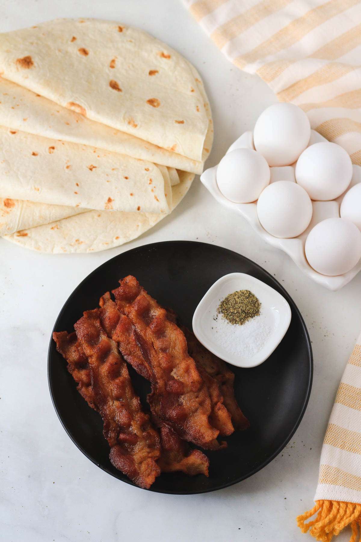 A white counter with ingredients to make bacon and egg breakfast burritos for the freezer.