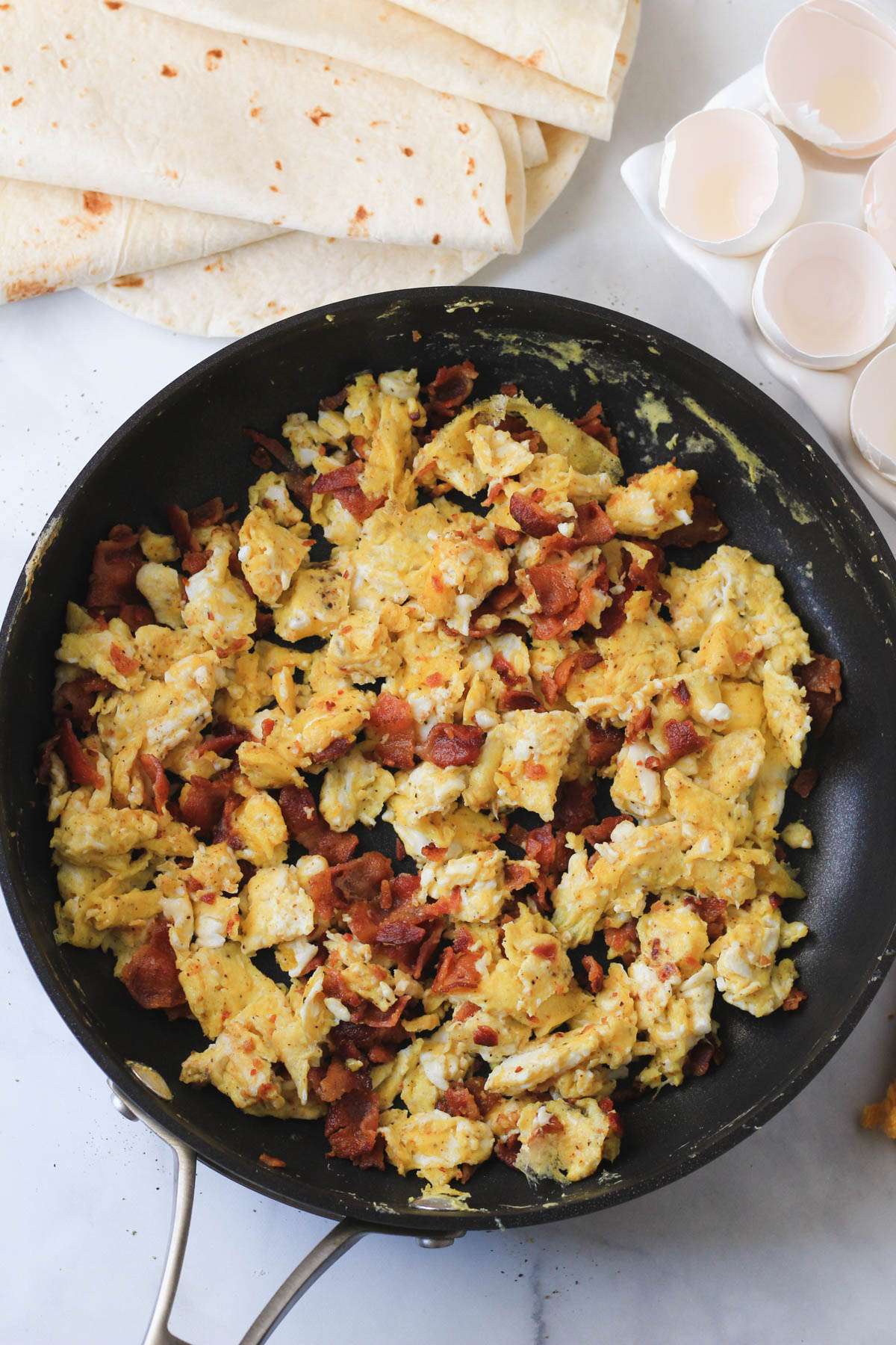 A non-stick skillet with bacon and egg cooked together and a pile of tortillas in the back left.