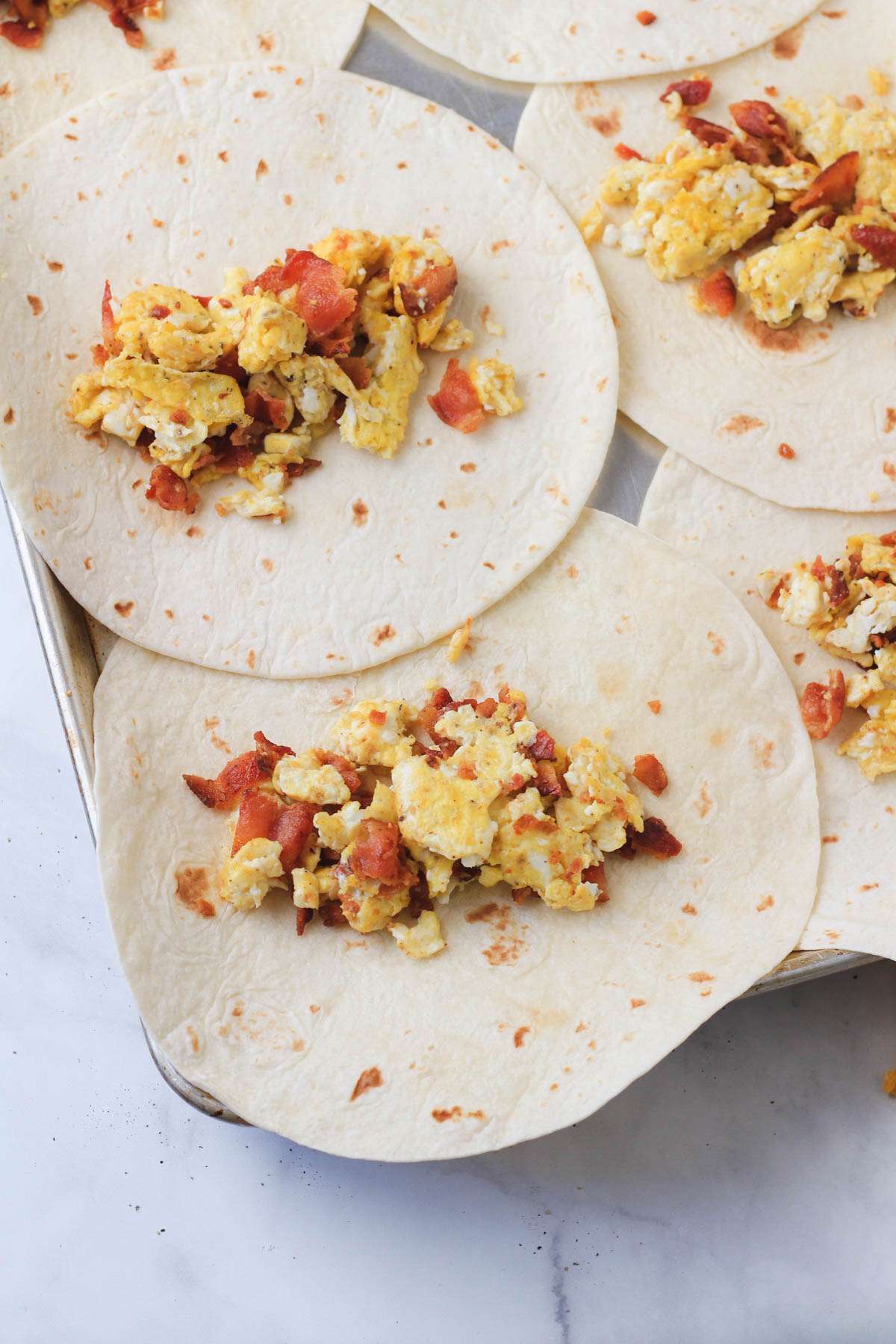 A rimmed baking sheet topped with flour tortillas and filled with bacon and egg burrito mixture.