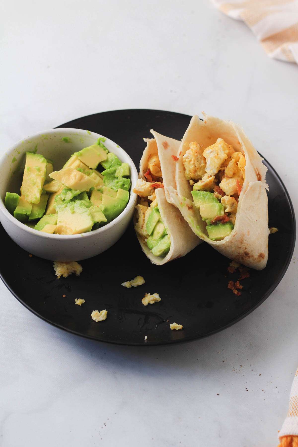 A black plate with a sliced bacon and egg breakfast burrito next to a white bowl with fresh sliced avocado.