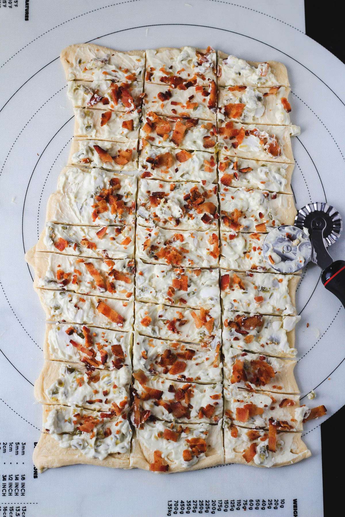 A pastry mat with a pastry cutter on the right side showing how to cut the crescent roll dough topped with jalapeño cream cheese and cooked bacon.