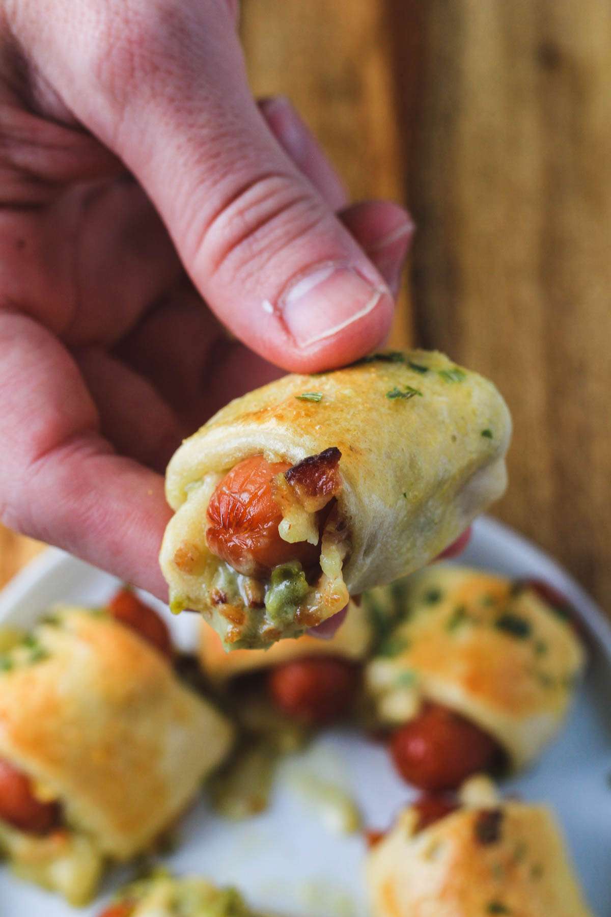 A hand holding a jalapeño popper pig in a blanket over a plate of other pigs in a blanket.