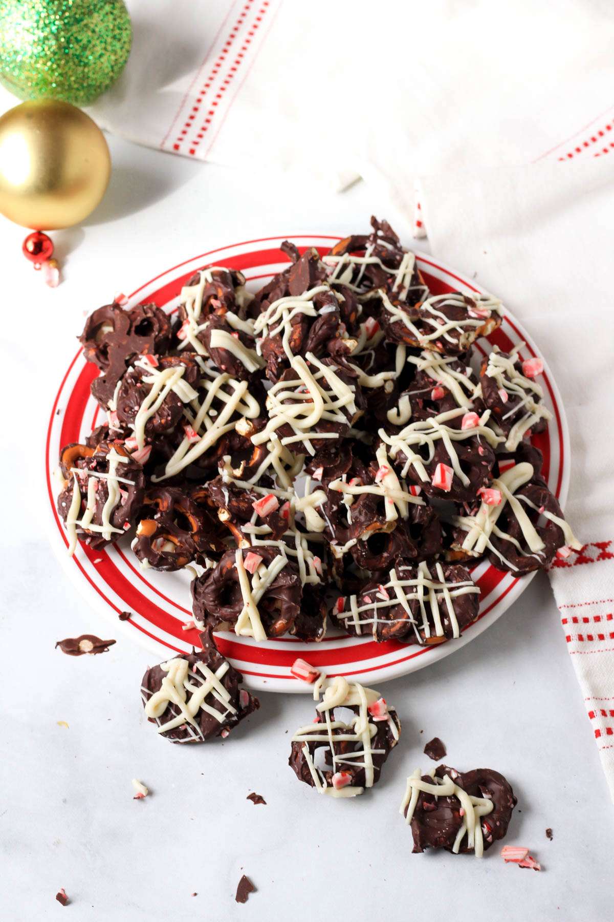 A red rimmed plate topped with a pile of peppermint bark pretzels on a white counter with two ornaments in the back left.