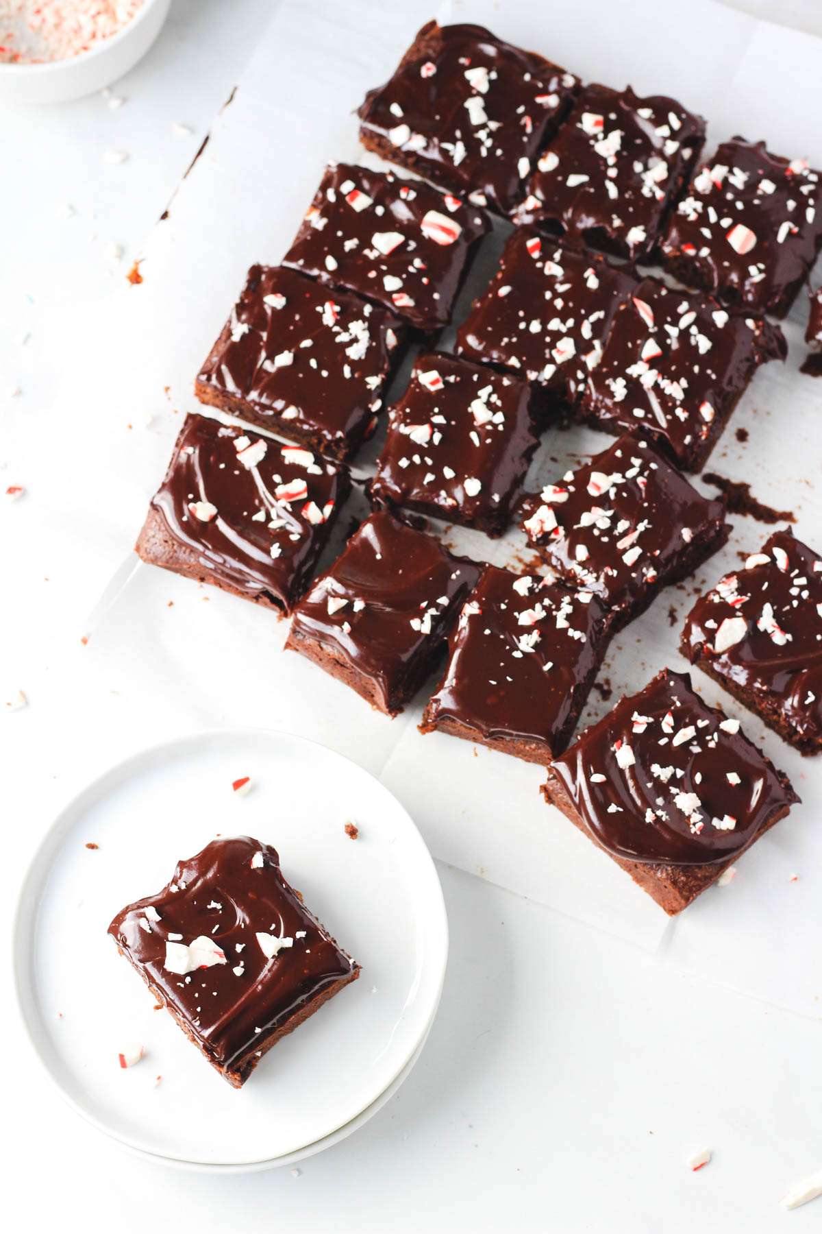 A small white plate with one peppermint brownie in the bottom left with a parchment paper of sliced peppermint brownies in the back right.