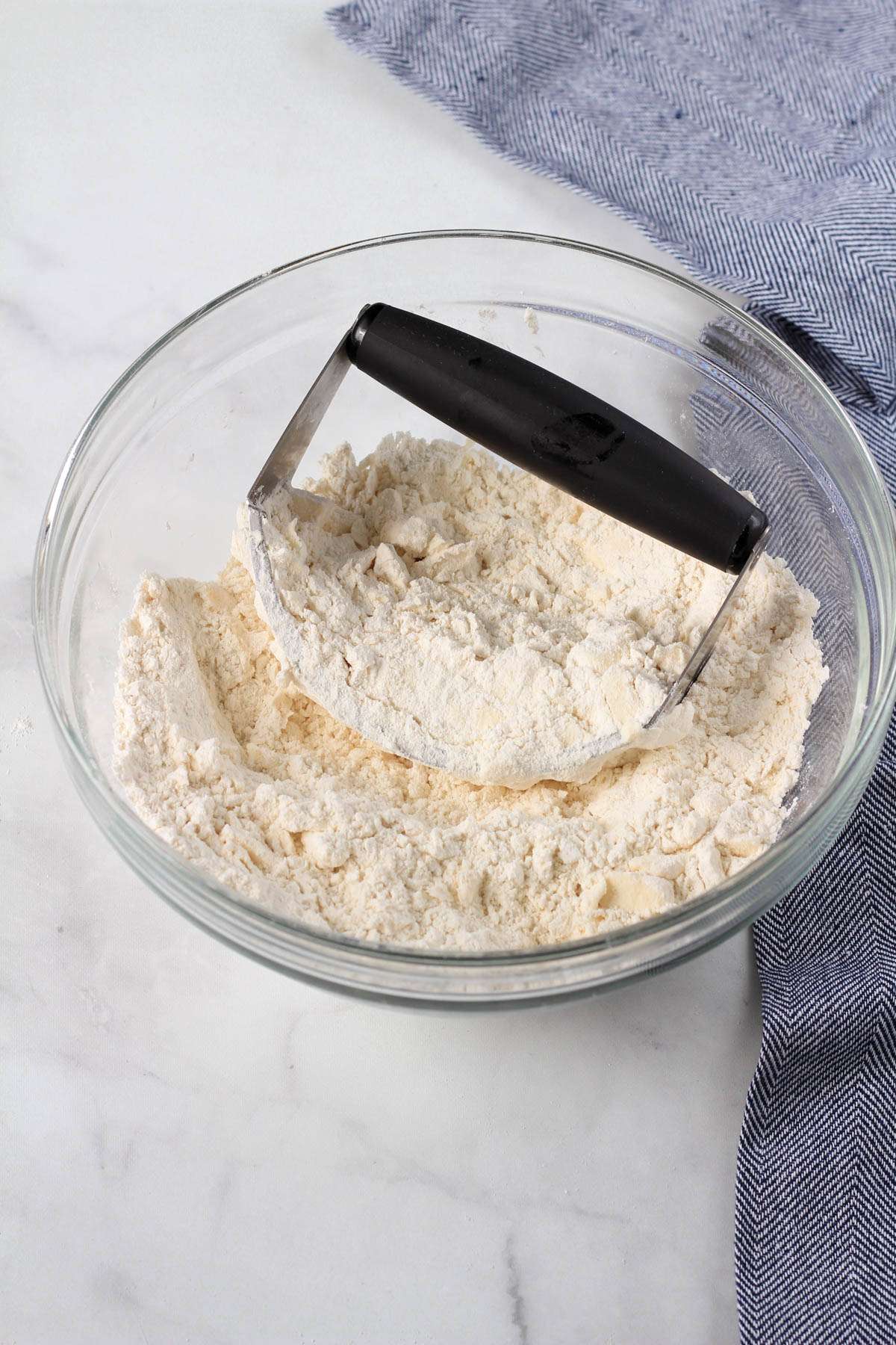 A glass mixing bowl with a pastry cutter cutting the vegan butter into the flour.
