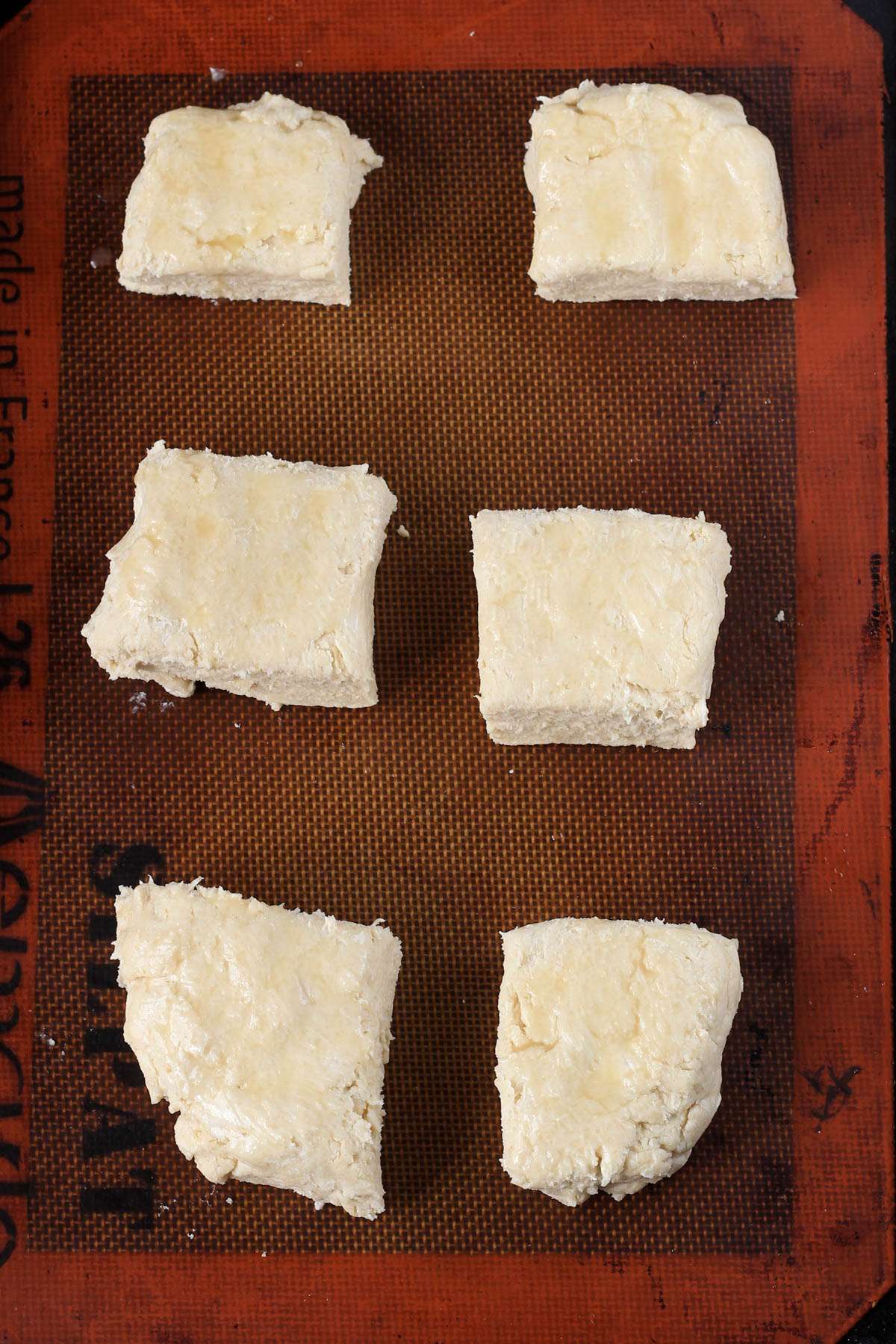 A pastry mat lined baking sheet with uncooked buttermilk biscuits brushed with vegan butter.