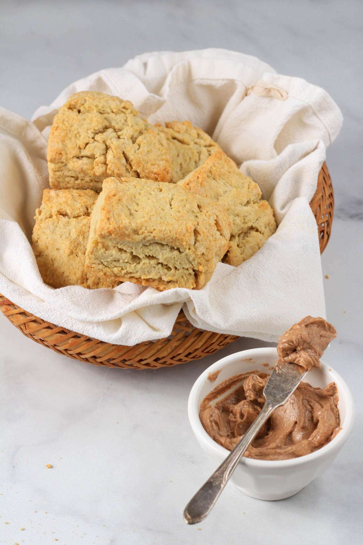 A brown basket with a white towel filled with dairy-free biscuits with a bowl of cinnamon honey butter in the front.