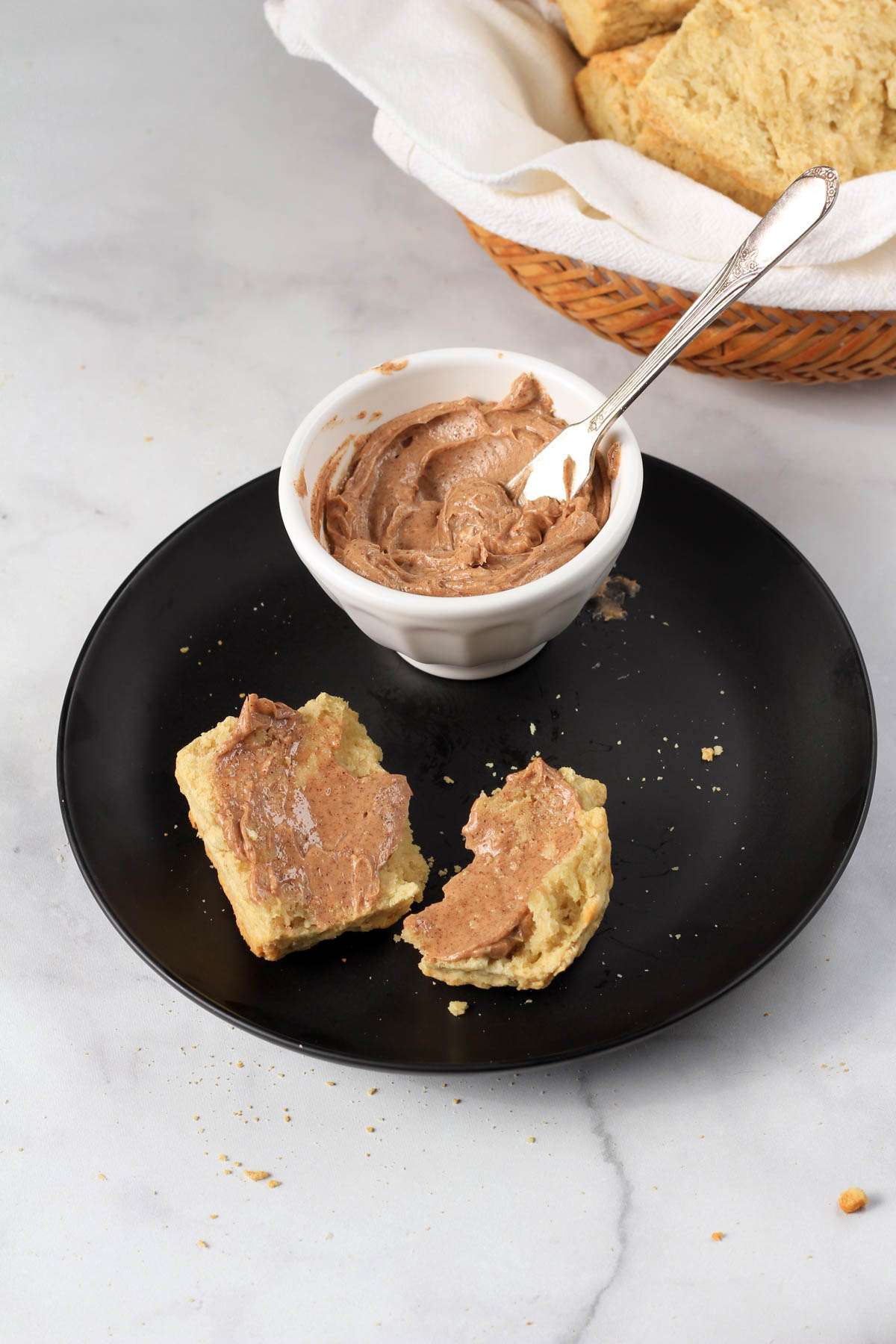 A black plate with half of a biscuit topped with cinnamon honey butter and a white bowl with a butter knife dipped in the cinnamon honey butter.