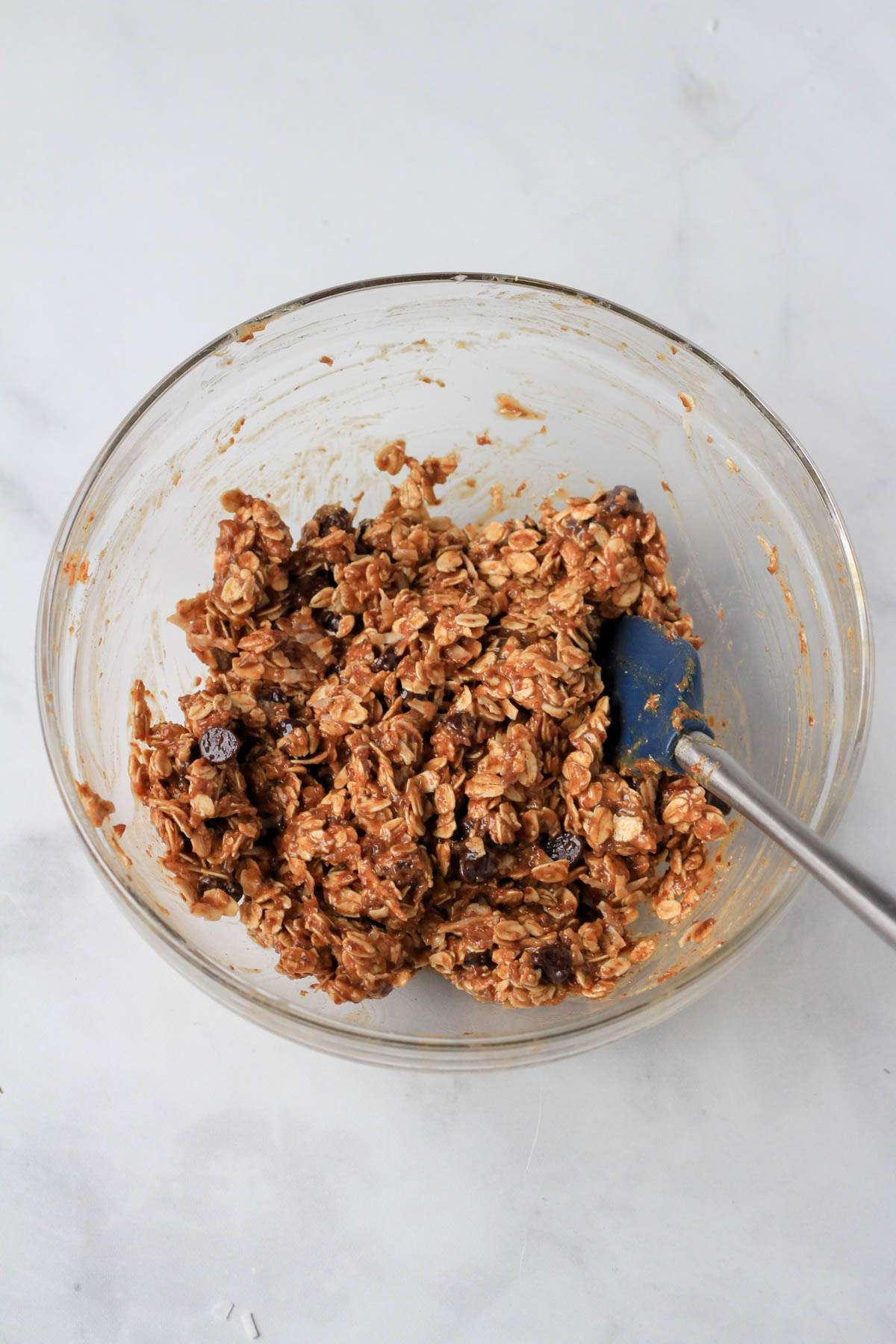 A glass bowl with a blue and silver spatula and the almond joy lactation bites mixed ingredients.