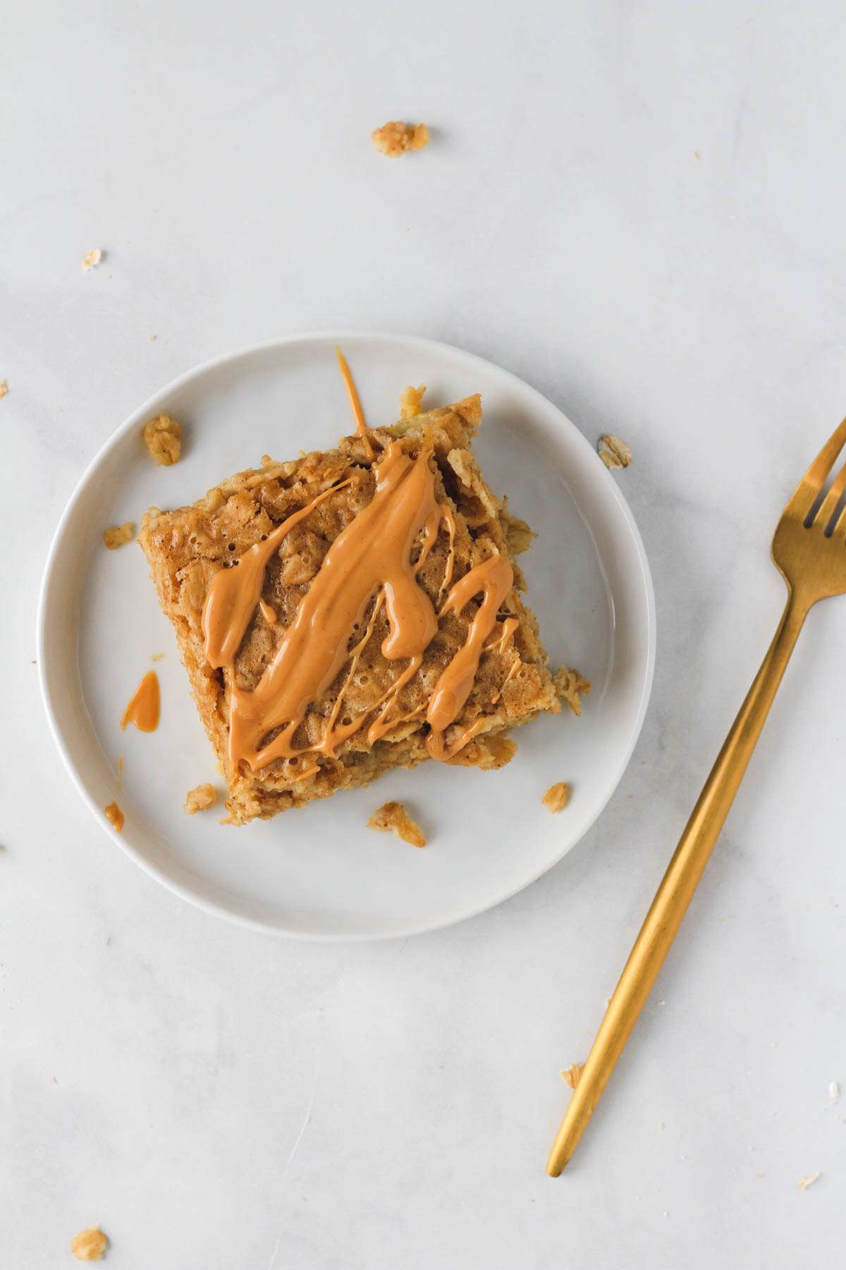 A small white plate with a peanut butter baked oatmeal bar drizzled with peanut butter and a gold fork to the right.