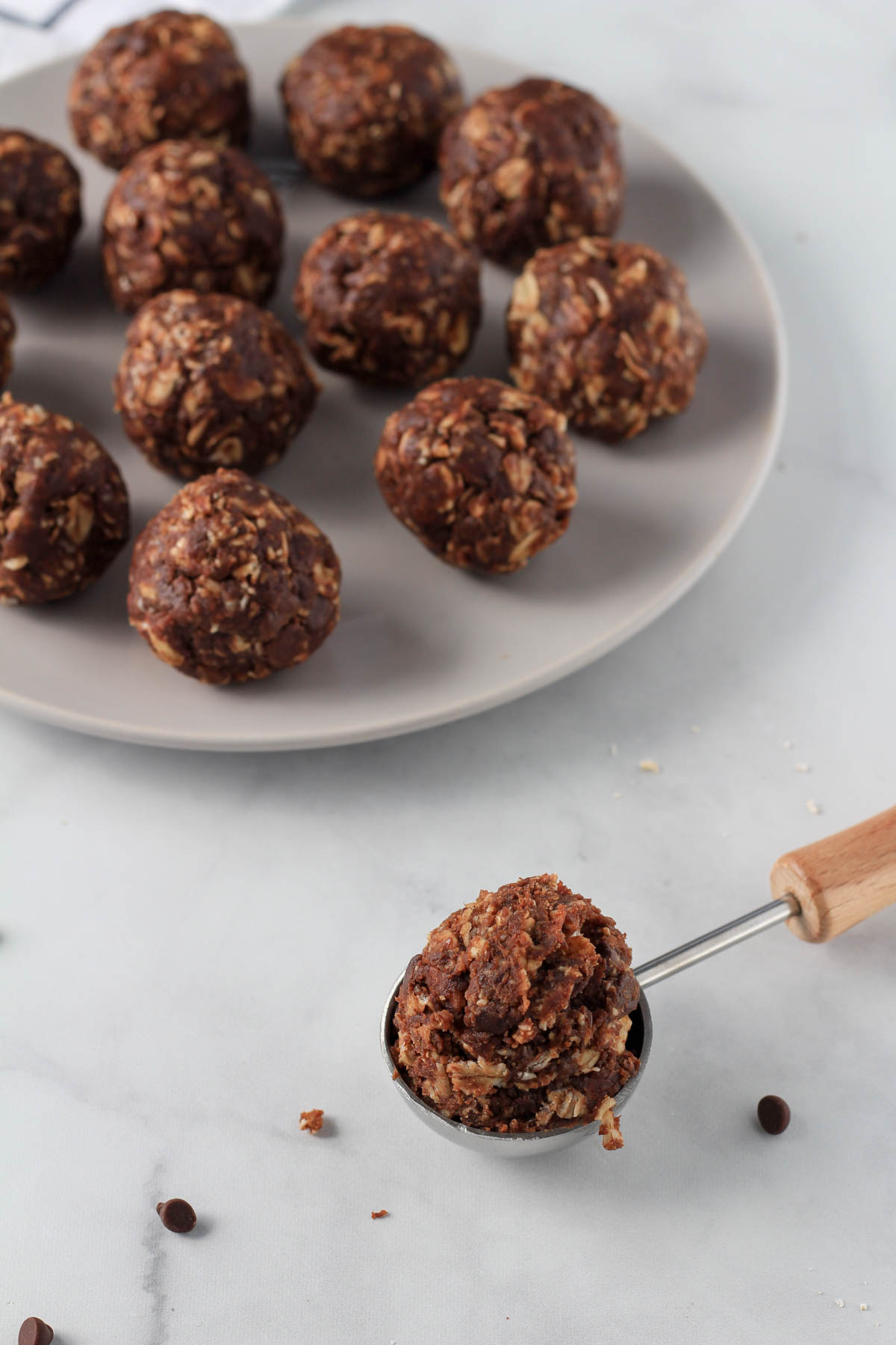A wooden handled scoop of chocolate almond lactation bite dough in front of a plate of rolled balls.