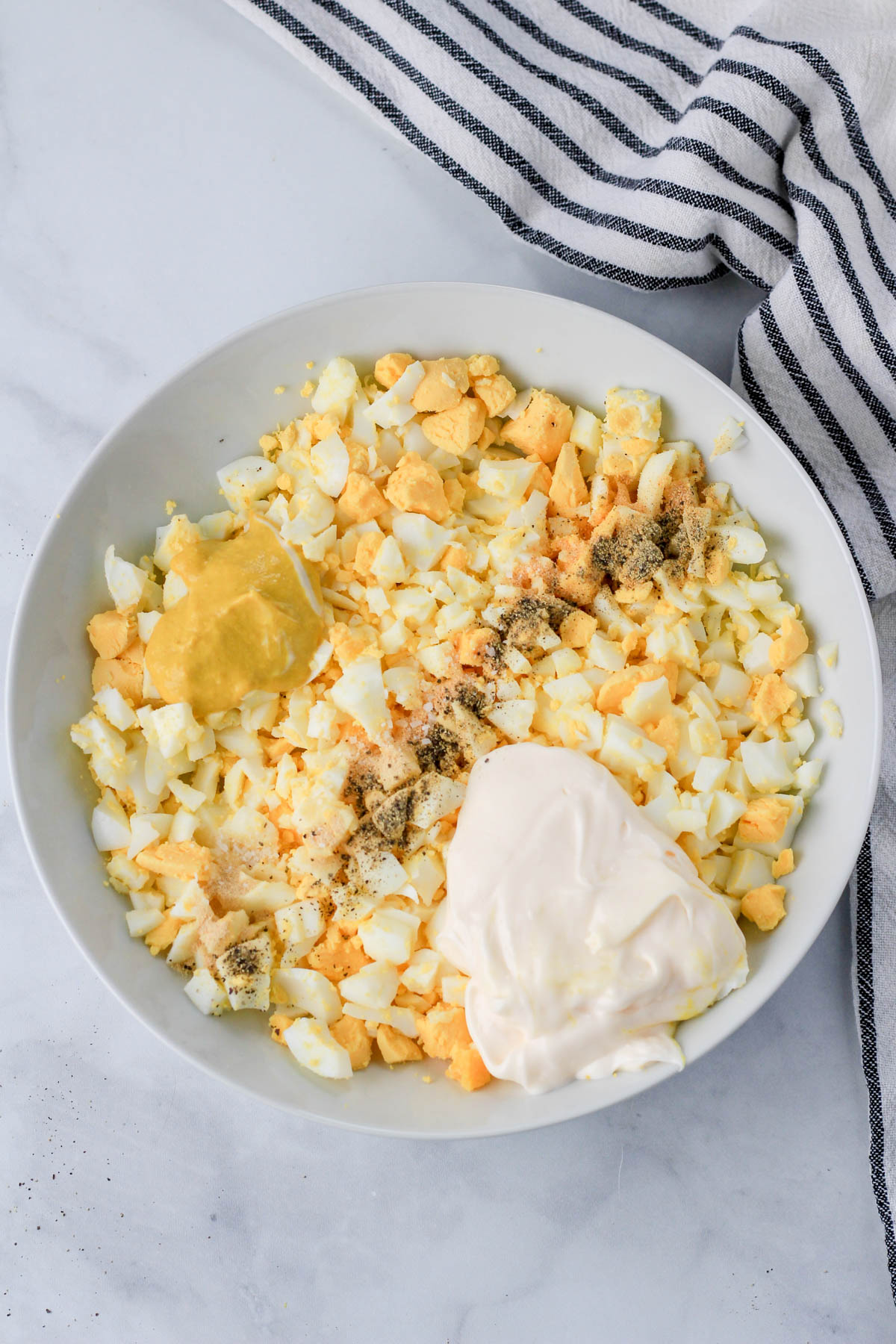 A white bowl with the ingredients for egg salad before mixing on a white counter with a blue and white striped dish towel.