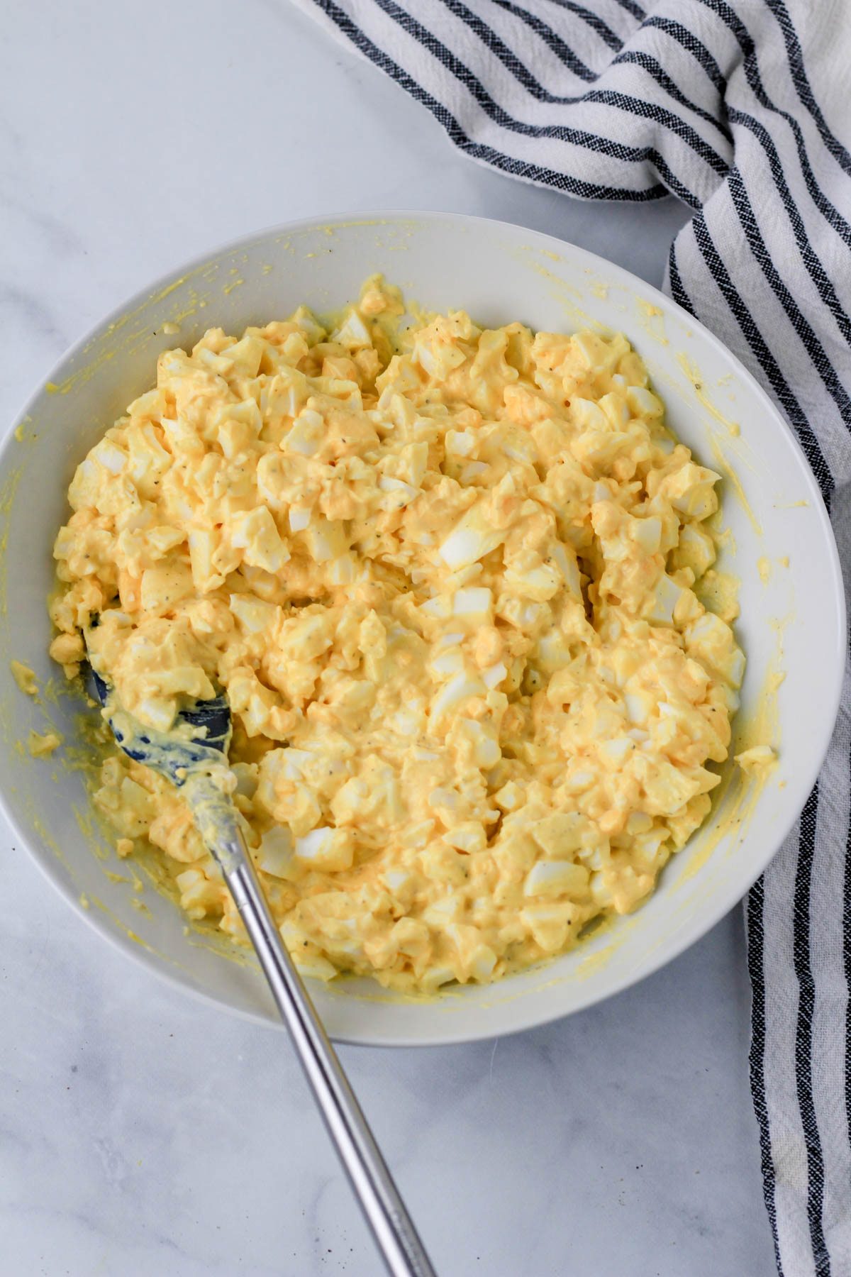 A small white bowl with mixed egg salad and a blue and silver spatula to the left side in the bowl.