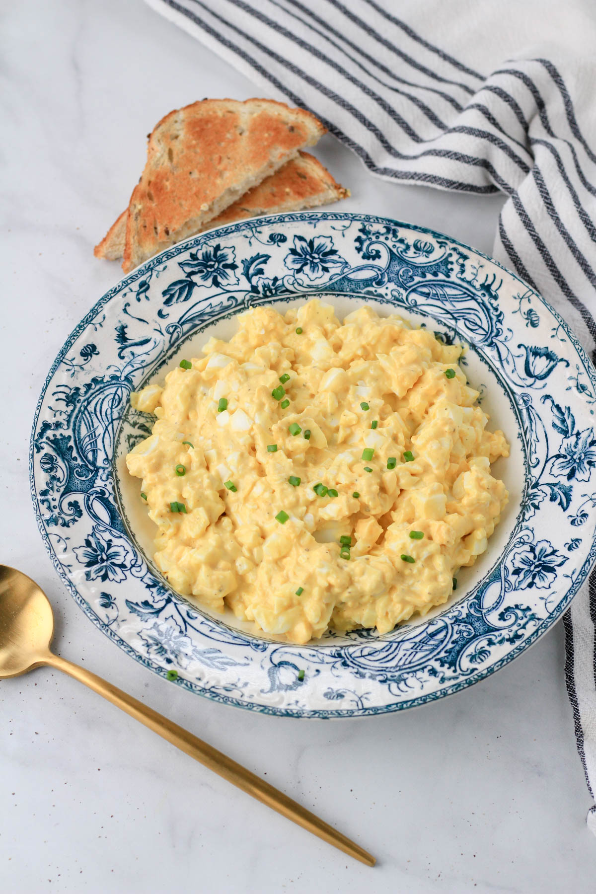 A blue and white rimmed bowl with classic egg salad and toasted bread in the back.