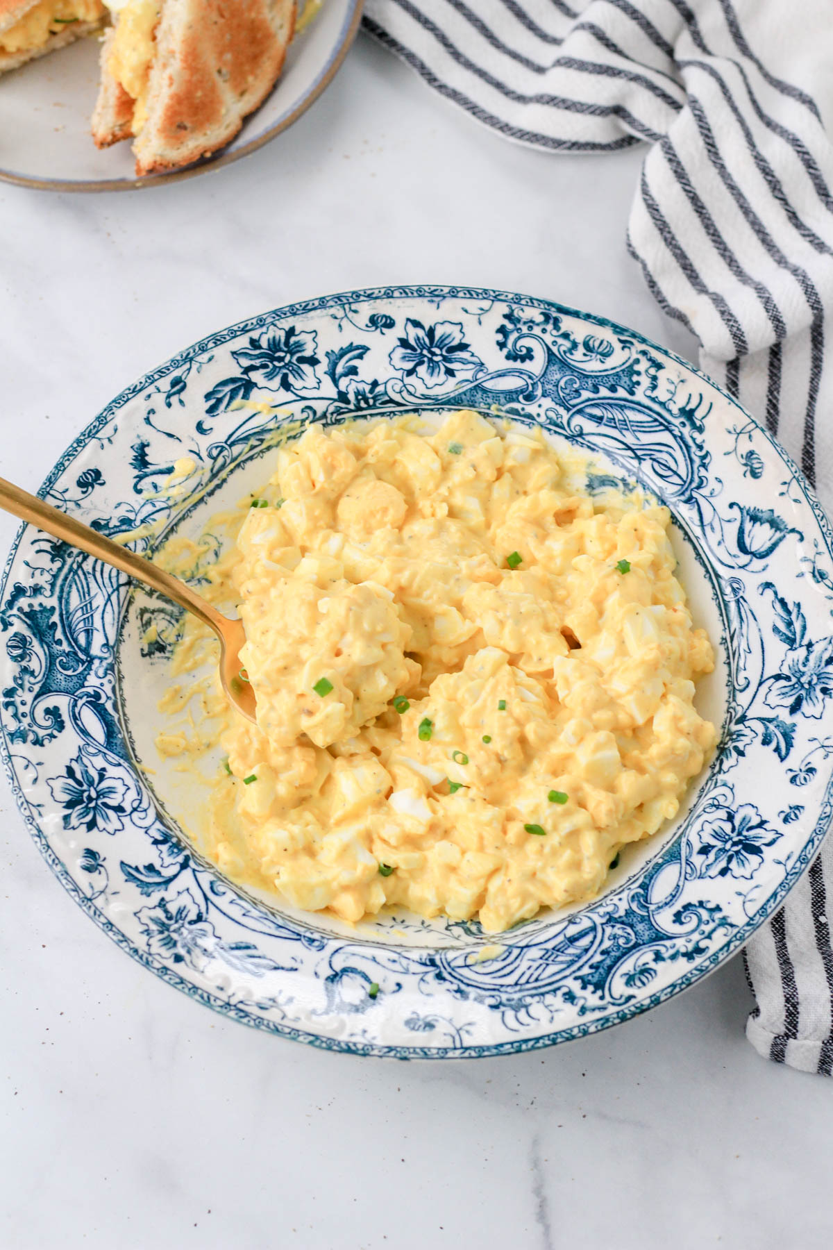 A serving bowl of egg salad with a gold spoon in the left side of the bowl and a toasted sandwich in the back left corner.