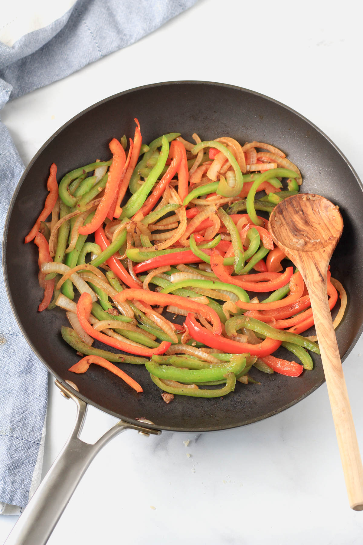 A non-stick skillet with bell peppers and onion with a wooden spoon to the right.