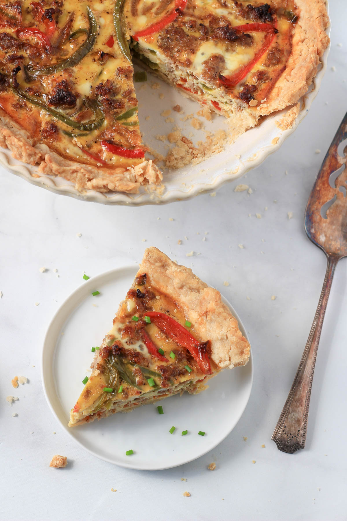 A small white plate with a slice of sausage and peppers quiche in front and a pie plate with the rest of the quiche and a pie server to the right.