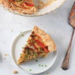 A small white plate with a slice of sausage and peppers quiche with a silver pie server to the right.
