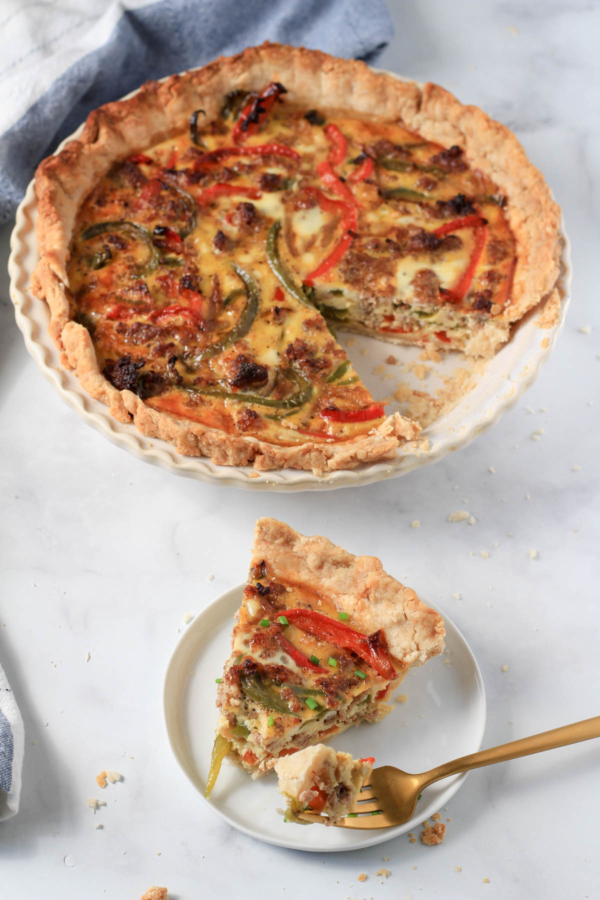 A small white plate with a slice of sausage and bell pepper quiche with a bite on a gold fork and the rest of the quiche in the back.